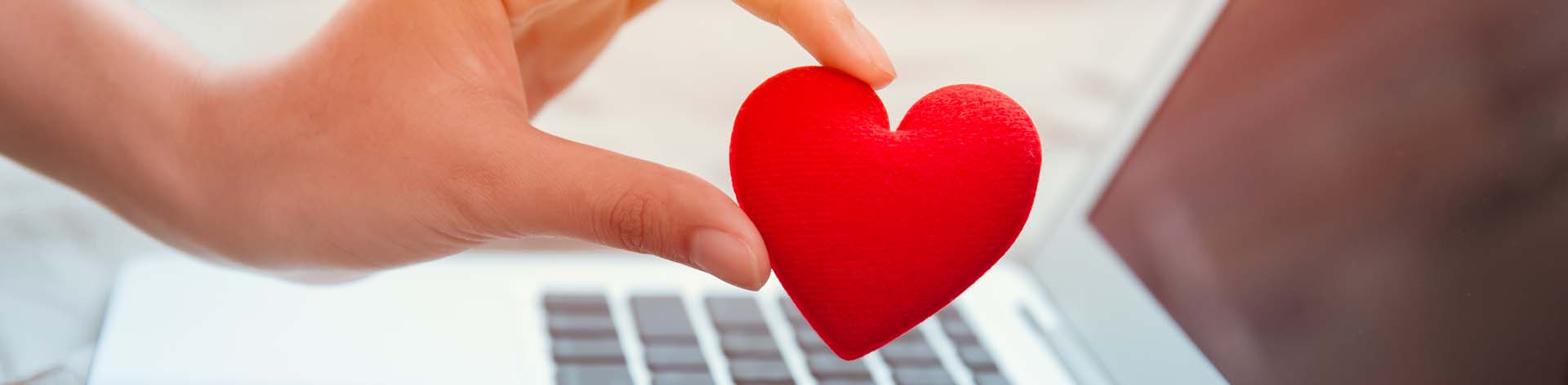 A hand holds a red fabric heart over a laptop.